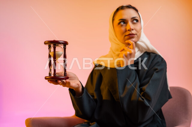 مفهوم أداره الاعمال والاوقات، بورتريه لسيده عربيه خليجيه عمانيه محجبه تجلس على كرسي مريح تحمل بيدها الساعة الرملية الزجاجية (البنكام) بايماءات اهميه مراقبة الوقت, تحديد مواعيد العمل وإدارة الجدول الزمني، خلفيه ملونه