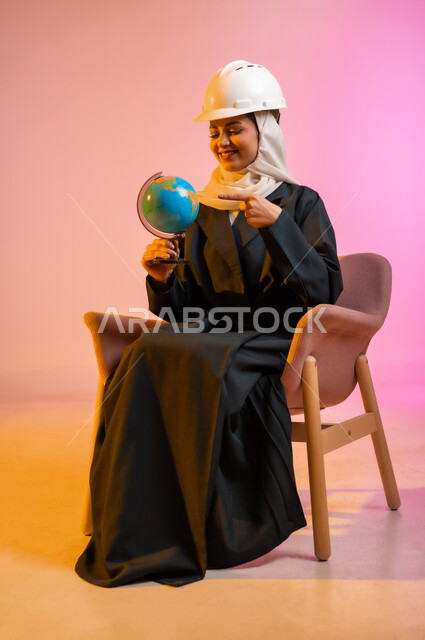 مفهوم البحث والاستكشاف حول العالم، بورتريه لسيده عربيه خليجيه عماني مبتسمه ترتدي العباءه والحجاب تجلس على كرسي مريح وتضع فوق رأسها خوذه المهندس تحمل بيدها مجسما للكرة الأرضية وتشير اليه بيدها الى مكان ما، مفهوم السياحه والسفر واستكشاف العالم، خلفيه ملونه