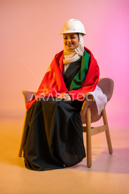 المشاركة بالاعياد والمناسبات الوطنية، الاحتفال بذكرى اليوم الوطني العماني، الاعتزاز والفخر بالرموز والراية العمانية، بورتريه لشابه عربيه خليجيه عمانيه ترتدي العباءه والحجاب وخوذه الحمايه تجلس على كرسي مريح وتلف حول جسدها العلم العماني ، مفهوم حب الوطن، خلفية ملونه