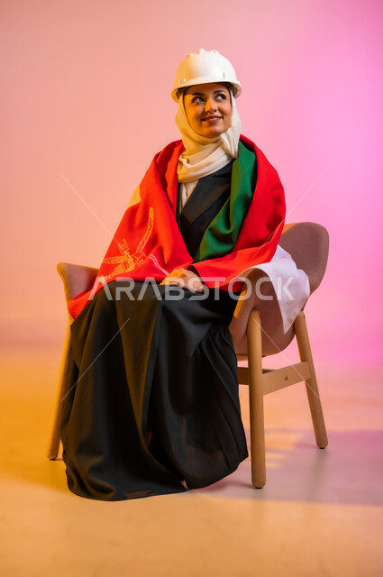 المشاركة بالاعياد والمناسبات الوطنية، الاحتفال بذكرى اليوم الوطني العماني، الاعتزاز والفخر بالرموز والراية العمانية، بورتريه لشابه عربيه خليجيه عمانيه ترتدي العباءه والحجاب وخوذه الحمايه تجلس على كرسي مريح وتلف حول جسدها العلم العماني ، مفهوم حب الوطن، خلفية ملونه