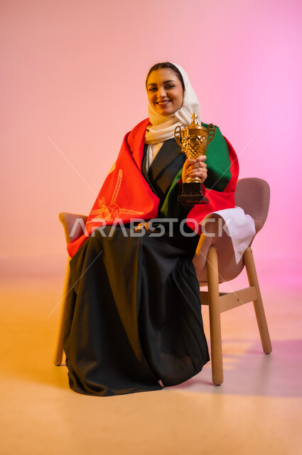 النظر الى الكاميرا بايماءات السعادة والسرور، الاعتزاز والفخر بالانتماء للهوية، الاحتفال بذكرى اليوم الوطني، بورتريه لشابه عربيه خليجيه عمانيه محجبة ترتدي العباءه السوداء تضع العلم على اكتافها تحمل بيدها كأس العالم الذهبي لرياضة كرة القدم، مفهوم الربح والفوز، خلفية ملونه
