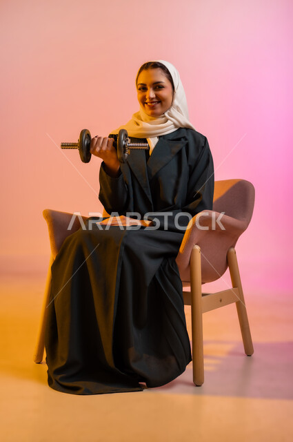 ممارسة التمارين الرياضية ورفع الاثقال مع الدمبل، بورتريه لشابة عربية خليجية عمانية محجبة مبتسمة ترتدي العباءة السوداء تجلس على الكرسي المريح تحمل بيدها الأوزان الحديدية الثقيلة، المحافظة على الصحة واللياقة الجسدية والرشاقة البدنية، خلفية ملونة
