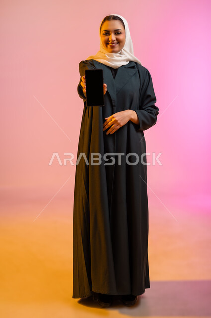 Having a good time exchanging news and conversations remotely, portrait of a veiled Gulf Arab Omani woman holding a mobile phone in her hand and pointing at it with a blank black screen, facial gestures indicating happiness and pleasure, colorful background