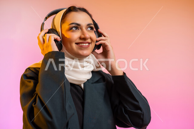 استخدام التكنولوجيا والأجهزة التقنية الحديثة المتطورة, بورتريه لفتاة عربية خليجية عمانيه محجبة ترتدي العباءة تضع سماعة الهيدفون على اذنها وتشير اليها بايماءات الانصات والاستماع بسعاده وسرور, الاستمتاع بسماع الموسيقى من خلال السماعه, خلفية ملونه