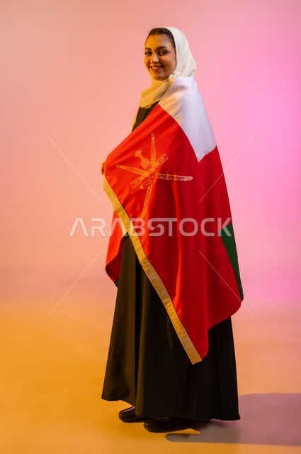 Pride and pride in belonging to the identity, participation in national holidays and occasions, celebration of the Omani National Day, pride and pride in the Omani symbols and flag, portrait of a young Arab Gulf Omani woman wearing an abaya and hijab and wrapping the Omani flag around her body, the concept of patriotism, colorful background