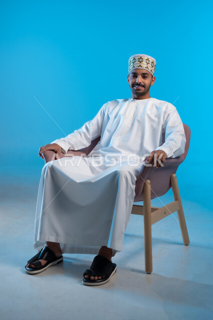 الاسترخاء والاستمتاع بالأجواء الهادئة، بورتريه لرجل عربي خليجي عماني يرتدي الدشداشه والكمه يجلس على كرسي مريح ينظر الى الكاميرا بايماءات السعادة والسرور، مفهوم الاناقة والاهتمام بالمظهر الخارجي, خلفية زرقاء