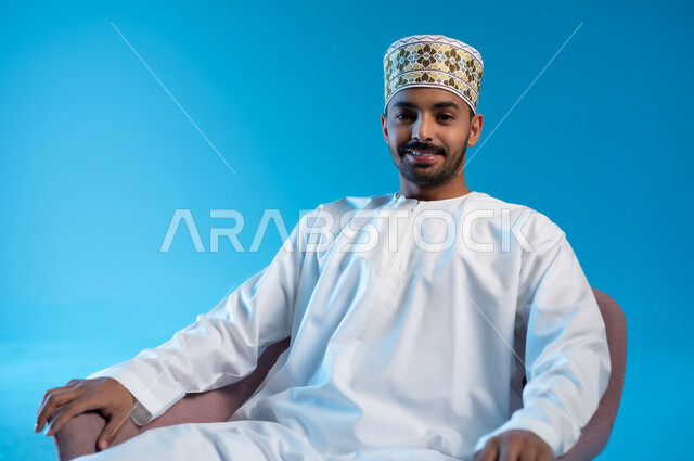 مفهوم الاناقة والاهتمام بالمظهر الخارجي, الاسترخاء والاستمتاع بالأجواء الهادئة، بورتريه لرجل عربي خليجي عماني يرتدي الدشداشه والكمه يجلس على كرسي مريح ينظر الى الكاميرا بايماءات السعادة والسرور، خلفية زرقاء