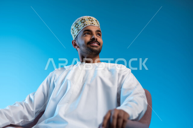 الاستمتاع و الاسترخاء  بالأجواء الهادئة، مفهوم الاناقة والاهتمام بالمظهر الخارجي, بورتريه مقرب لرجل عربي خليجي عماني يرتدي الدشداشه والكمه يجلس على كرسي مريح ينظر الى شيء ما بايماءات السعادة والسرور، خلفية زرقاء