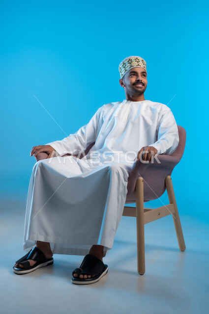 الاسترخاء والاستمتاع بالأجواء الهادئة، مفهوم الاناقة والاهتمام بالمظهر الخارجي, بورتريه لرجل عربي خليجي عماني يرتدي الدشداشه والكمه يجلس على كرسي مريح ينظر الى شيء ما بايماءات السعادة والسرور، خلفية زرقاء