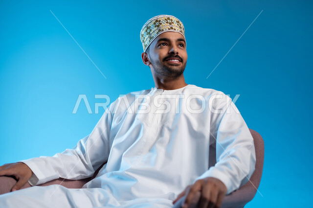 مفهوم الاناقة والاهتمام بالمظهر الخارجي, الاستمتاع و الاسترخاء  بالأجواء الهادئة، بورتريه لرجل عربي خليجي عماني يرتدي الدشداشه والكمه يجلس على كرسي مريح ينظر الى شيء ما بايماءات السعادة والسرور، خلفية زرقاء