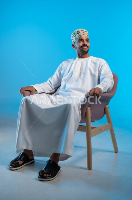 مفهوم الاناقة والاهتمام بالمظهر الخارجي, الاستمتاع و الاسترخاء  بالأجواء الهادئة، بورتريه لرجل عربي خليجي عماني يرتدي الدشداشه والكمه يجلس على كرسي مريح ينظر الى شيء ما بايماءات السعادة والسرور، خلفية زرقاء