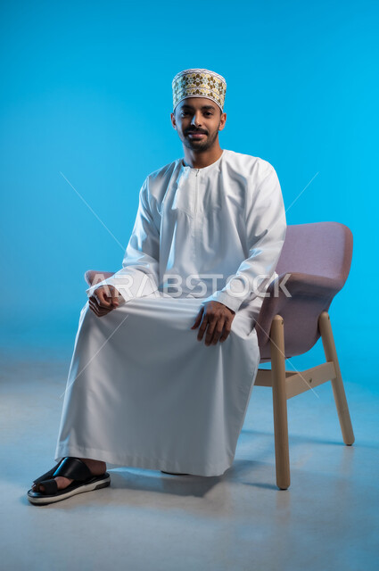 الاسترخاء والاستمتاع بالأجواء الهادئة، بورتريه لرجل عربي خليجي عماني يرتدي الدشداشه والكمه يجلس على كرسي مريح ينظر الى الكاميرا بايماءات السعادة والسرور، مفهوم الاناقة والاهتمام بالمظهر الخارجي, خلفية زرقاء