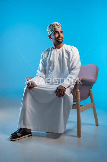 الاسترخاء والاستمتاع بالأجواء الهادئة، مفهوم الاناقة والاهتمام بالمظهر الخارجي, بورتريه لرجل عربي خليجي عماني يرتدي الدشداشه والكمه يجلس على كرسي مريح ينظر الى شيء ما بايماءات السعادة والسرور، خلفية زرقاء