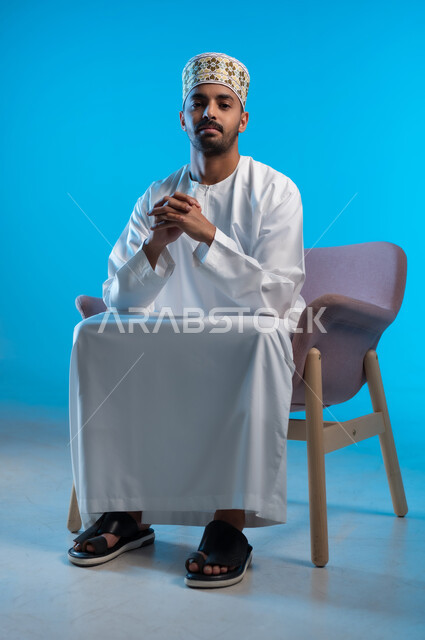 The concept of elegance and interest in the external appearance, relaxation and enjoyment of the calm atmosphere, a portrait of an Arab Gulf Omani man wearing a dishdasha and a cloak sitting on a comfortable chair with crossed hands looking at the camera with gestures of happiness and pleasure, blue background