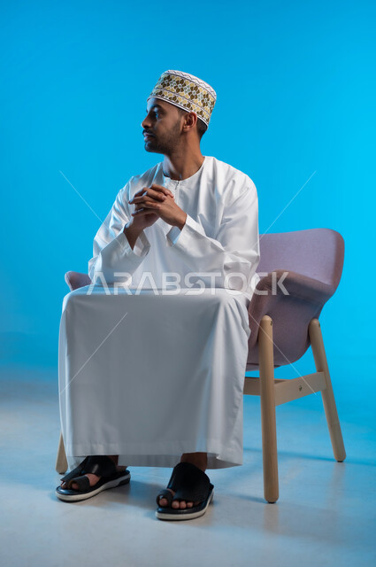 مفهوم الاناقة والاهتمام بالمظهر الخارجي, الاستمتاع و الاسترخاء  بالأجواء الهادئة، بورتريه لرجل عربي خليجي عماني يرتدي الدشداشه والكمه يجلس على كرسي مريح ينظر الى شيء ما بايماءات السعادة والسرور، خلفية زرقاء