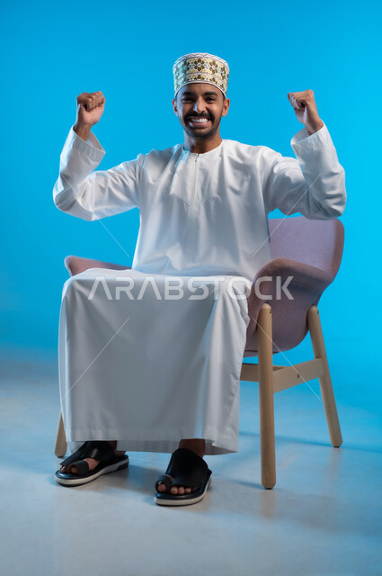 مفهوم الاناقة والاهتمام بالمظهر الخارجي, الاستمتاع و الاسترخاء بالأجواء الهادئة، بورتريه لرجل عربي خليجي عماني يرتدي الدشداشه والكمه يجلس على كرسي مريح يرفع قبضة يده بتعابير الفوز و الربح، خلفية زرقاء