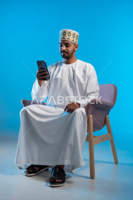 تصفح مواقع التواصل الاجتماعي, التواصل والدردشة مع الأهل والأصدقاء عبر تطبيقات الجوال, بورتريه لشاب عربي خليجي عماني يرتدي الكمة و الدشداشة يجلس على كرسي مريح يحمل الهاتف المحمول, استخدام التقنيات الحديثة المتطورة, خلفية زرقاء