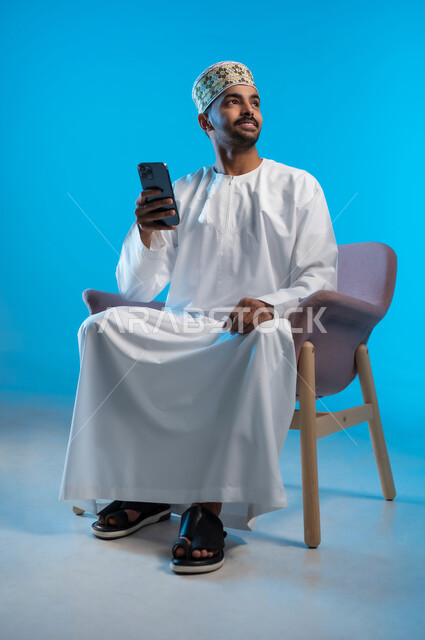 تصفح مواقع التواصل الاجتماعي, التواصل والدردشة مع الأهل والأصدقاء عبر تطبيقات الجوال, بورتريه لشاب عربي خليجي عماني يرتدي الكمة و الدشداشة يجلس على كرسي مريح يحمل الهاتف المحمول, استخدام التقنيات الحديثة المتطورة, خلفية زرقاء