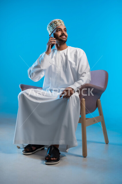 التواصل مع الاهل و الاصدقاء, ايماءات السعادة و السرور، بورتريه لرجل عربي خليجي عماني يرتدي الدشداشه و الكمه يجلس على كرسي مريح يقوم بإجراء مكالمة هاتفية عبر الهاتف المحمول، استخدام التقنيات الحديثة المتطورة, خلفية زرقاء