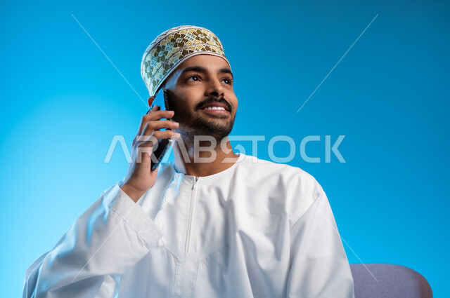 ايماءات السعادة و السرور، التواصل مع الاهل و الاصدقاء, بورتريه مقرب لرجل عربي خليجي عماني يرتدي الدشداشه و الكمه يجلس على كرسي مريح يقوم بإجراء مكالمة هاتفية عبر الهاتف المحمول، استخدام التقنيات الحديثة المتطورة, خلفية زرقاء