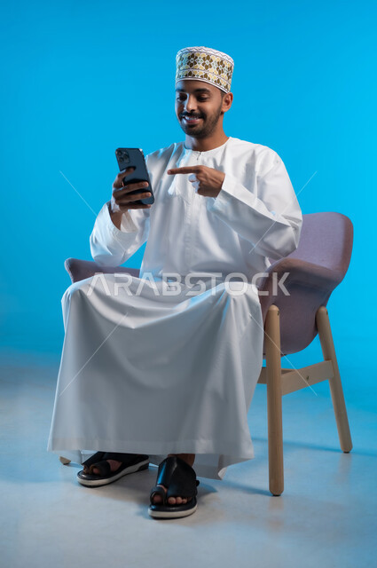 استخدام التقنيات الحديثة المتطورة, تصفح مواقع التواصل الاجتماعي, التواصل والدردشة مع الأهل والأصدقاء عبر تطبيقات الجوال, بورتريه لشاب عربي خليجي عماني يرتدي الكمة و الدشداشة يجلس على كرسي مريح يشير باصبع السبابة الى الهاتف المحمول, خلفية زرقاء