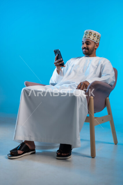 Browsing social networking sites, communicating and chatting with family and friends via mobile applications, portrait of a young Gulf Arab Omani man wearing a turban and a dishdasha sitting on a comfortable chair using a mobile phone, using modern advanced technologies, blue background