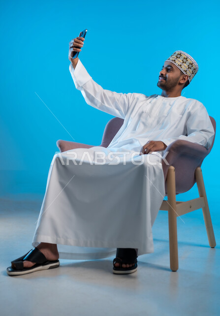 توثيق اللحظات و الذكريات, بورتريه لشاب عربي  خليجي عماني يرتدي الكمة و الدشداشة يجلس على كرسي مريح يقوم بالتقاط صور سيلفي من خلال الهاتف المحمول, خلفية زرقاء
