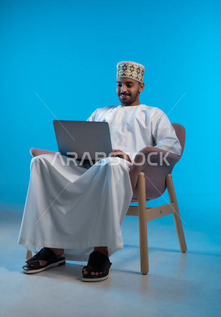 إدارة شؤون الأعمال وتنظيمها، استخدام التقنيات الحديثة المتطورة، بورتريه لشاب عربي خليجي عماني يرتدي الكمة و الدشداشة يجلس على كرسي مريح يعمل عبر الحاسب المحمول, ايماءات التركيز و التمعن, خلفية زرقاء