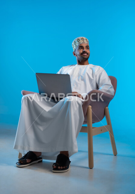 إدارة شؤون الأعمال وتنظيمها، استخدام التقنيات الحديثة المتطورة، بورتريه لشاب عربي خليجي عماني يرتدي الكمة و الدشداشة يجلس على كرسي مريح ينظر الى شيء ما  يعمل عبر الحاسب المحمول, خلفية زرقاء