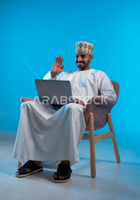 رفع كف اليد للاعلى بإيماءات القاء التحية و السلام, عقد إجتماع عمل عبر الإنترنت، مناقشة إستراتيجية المشروع, إدارة شؤون الأعمال وتنظيمها، استخدام التقنيات الحديثة المتطورة، بورتريه لشاب عربي خليجي عماني يرتدي الكمة و الدشداشة يجلس على كرسي مريح يقوم بإجراء مكالمة فيديو من خلال الحاسب المحمول, خلفية زرقاء