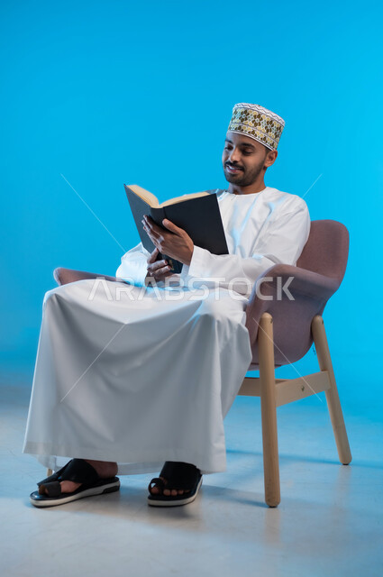 ممارسة هواية المطالعة, مفهوم الشغف بالقراءة, بورتريه لشاب عربي خليجي عماني يرتدي الكمة والدشداشة يجلس على كرسي مريح و يقوم بقراءه كتاب, كتب وروايات هادفه ومسليه ، التعلم والتثقيف من خلال قراءه الكتب الهادفه، خلفيه زرقاء