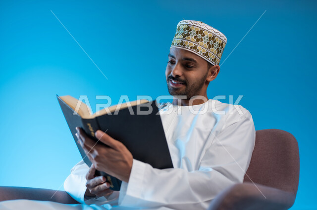 ممارسة هواية المطالعة, مفهوم الشغف بالقراءة, بورتريه لشاب عربي خليجي عماني يرتدي الكمة والدشداشة يجلس على كرسي مريح و يقوم بقراءه كتاب, كتب وروايات هادفه ومسليه ، التعلم والتثقيف من خلال قراءه الكتب الهادفه، خلفيه زرقاء