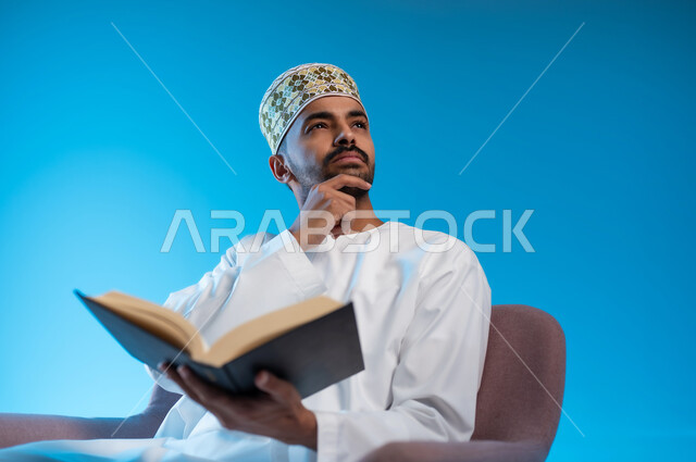 التعلم والتثقيف من خلال قراءه الكتب و الروايات الملية و الهادفه، ممارسة هواية المطالعة, بورتريه لشاب عربي خليجي عماني يرتدي الكمة و الدشداشة يجلس على كرسي مريح يمسك بيده كتاب يضع يده على ذقمه بإيماءات التفكير و الحيرة, خلفيه زرقاء