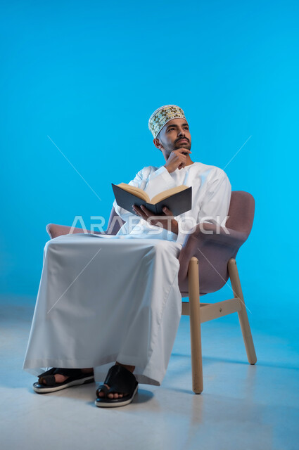 التعلم والتثقيف من خلال قراءه الكتب و الروايات الملية و الهادفه، ممارسة هواية المطالعة, بورتريه لشاب عربي خليجي عماني يرتدي الكمة و الدشداشة يجلس على كرسي مريح يمسك بيده كتاب يضع يده على ذقمه بإيماءات التفكير و الحيرة, خلفيه زرقاء