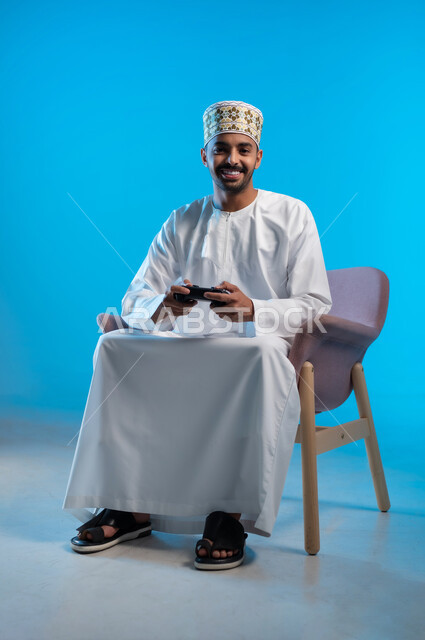 اجهزة وتقنيات الكترونية حديثة ترفيهيه, قضاء وقت ممتع ومسلي، التفاعل والاندماج مع ألعاب الفيديو والبلايستيشن، بورتريه لشاب عربي عماني خليجي يرتدي الكمة و الدشداشة يجلس على كرسي مريح ويحمل بيده عصا التحكم، خلفية زرقاء