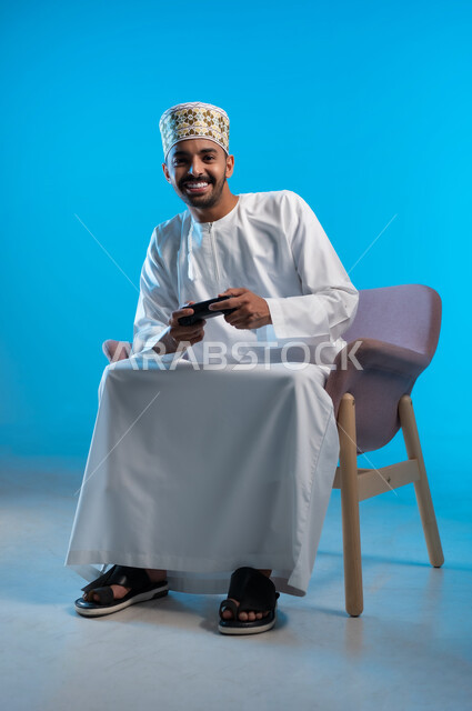 اجهزة وتقنيات الكترونية حديثة ترفيهيه, قضاء وقت ممتع ومسلي، التفاعل والاندماج مع ألعاب الفيديو والبلايستيشن، بورتريه لشاب عربي عماني خليجي يرتدي الكمة و الدشداشة يجلس على كرسي مريح ويحمل بيده عصا التحكم، خلفية زرقاء