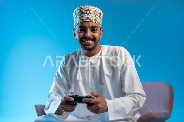 اجهزة وتقنيات الكترونية حديثة ترفيهيه, قضاء وقت ممتع ومسلي، التفاعل والاندماج مع ألعاب الفيديو والبلايستيشن، بورتريه لشاب عربي عماني خليجي يرتدي الكمة و الدشداشة يجلس على كرسي مريح ويحمل بيده عصا التحكم، خلفية زرقاء