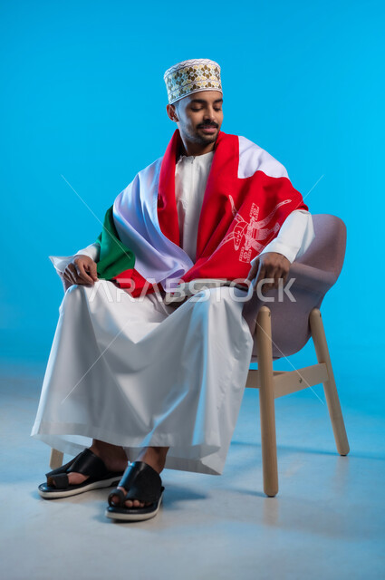 الاحتفال بذكرى اليوم الوطني العماني، إيماءات السرور و الفرح, الاعتزاز بالانتماء للهوية، الفخر بالرموز والراية العمانية، بورتريه لشاب عربي خليجي عماني يرتدي الكمة والدشداشة يجلس على الكرسي و يضع العلم على اكتافه، مفهوم حب الوطن، خلفية زرقاء