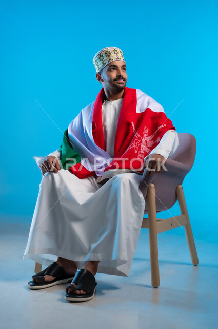 الاحتفال بذكرى اليوم الوطني العماني، إيماءات السرور و الفرح, الاعتزاز بالانتماء للهوية، الفخر بالرموز والراية العمانية، بورتريه لشاب عربي خليجي عماني يرتدي الكمة والدشداشة يجلس على الكرسي و يضع العلم على اكتافه، مفهوم حب الوطن، خلفية زرقاء