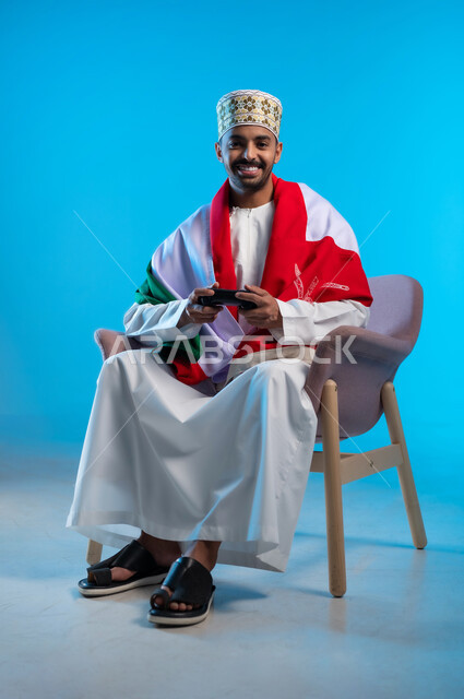 التفاعل والاندماج مع ألعاب البلايستيشن، الاحتفال بذكرى اليوم الوطني، الفخر بالانتماء للهوية، بورتريه لشاب عربي خليجي عماني يرتدي اللكمة و الدشداشة يجلس على كرسي مريح يضع العلم على اكتافه ويحمل بيده عصا التحكم، مفهوم الربح والفوز، خلفية زرقاء