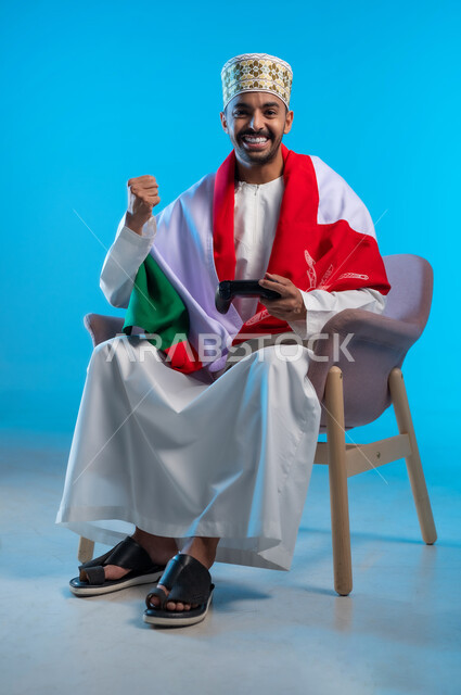الاحتفال بذكرى اليوم الوطني، التفاعل والاندماج مع ألعاب البلايستيشن، الفخر بالانتماء للهوية، بورتريه لشاب عربي خليجي عماني يرتدي اللكمة و الدشداشة يجلس على كرسي مريح يضع العلم على اكتافه ويحمل بيده عصا التحكم، رفع قبضة اليد بتعابير الربح والفوز، خلفية زرقاء