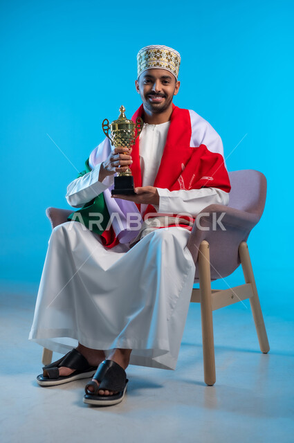 The concept of profit and victory, celebrating the National Day, pride and pride in belonging to the identity, a portrait of a young Gulf Arab Omani man wearing a lakma and a dishdasha sitting on a comfortable chair, placing the flag on his shoulders, holding the golden World Cup for football, blue background