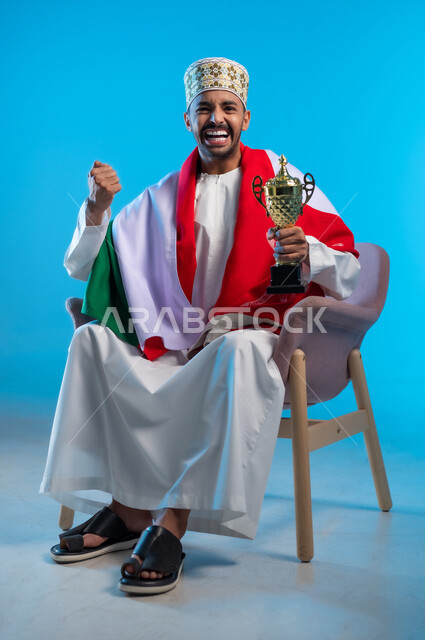 الاحتفال بذكرى اليوم الوطني، الاعتزاز والفخر بالانتماء للهوية، بورتريه لشاب عربي خليجي عماني يرتدي اللكمة و الدشداشة يجلس على كرسي مريح يضع العلم على اكتافه يحمل بيده كأس العالم الذهبي لرياضة كرة القدم، رفع قبضة اليد بتعابير الربح والفوز، خلفية زرقاء