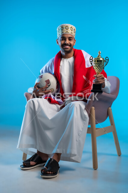 الاعتزاز والفخر بالانتماء للهوية، الاحتفال بذكرى اليوم الوطني، بورتريه لشاب عربي خليجي عماني يرتدي اللكمة و الدشداشة يجلس على كرسي مريح يضع العلم على اكتافه يحمل بيده كأس العالم الذهبي لرياضة كرة القدم، مفهوم الربح والفوز، خلفية ملونه