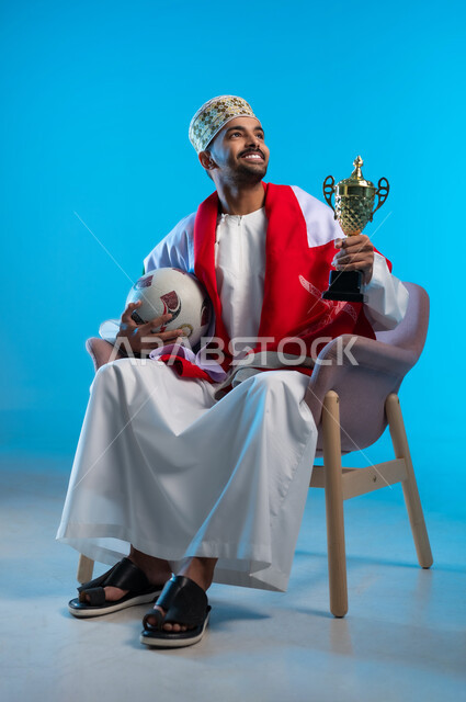 الاعتزاز والفخر بالانتماء للهوية، الاحتفال بذكرى اليوم الوطني، بورتريه لشاب عربي خليجي عماني يرتدي اللكمة و الدشداشة يجلس على كرسي مريح يضع العلم على اكتافه يحمل بيده كأس العالم الذهبي لرياضة كرة القدم، مفهوم الربح والفوز، خلفية ملونه
