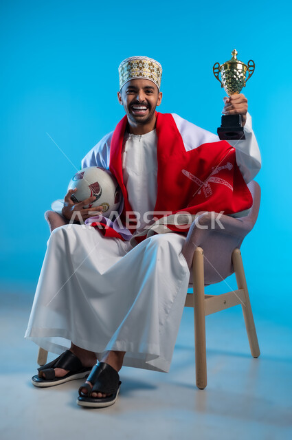 الاعتزاز والفخر بالانتماء للهوية، الاحتفال بذكرى اليوم الوطني، بورتريه لشاب عربي خليجي عماني يرتدي اللكمة و الدشداشة يجلس على كرسي مريح يضع العلم على اكتافه يحمل بيده كأس العالم الذهبي لرياضة كرة القدم، مفهوم الربح والفوز، خلفية ملونه