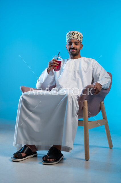 محاولة التخفيف من حرارة الصيف بالمشروب المنعش، الاستمتاع بالمشروبات المفيدة اللذيذة، بورتريه لرجل عربي خليجي عماني مبتسم يرتدي الكمة و الدشداشة يجلس على كرسي مريح ويستمتع بشرب العصير الطبيعي البارد, النظر الى الكاميرا بإيماءات السرور والسعادة, خلفية زرقاء