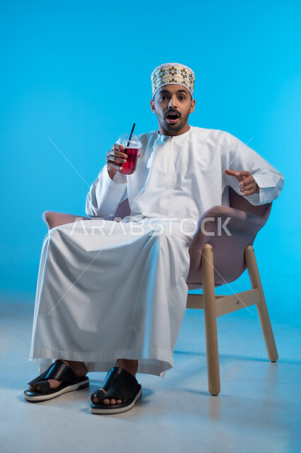 النظر الى الكاميرا بإيماءات المفاجئة و الذهول, محاولة التخفيف من حرارة الصيف بالمشروب المنعش، الاستمتاع بالمشروبات المفيدة اللذيذة، بورتريه لرجل عربي خليجي عماني مبتسم يرتدي الكمة و الدشداشة يجلس على كرسي مريح ويستمتع بشرب العصير الطبيعي البارد, خلفية زرقاء