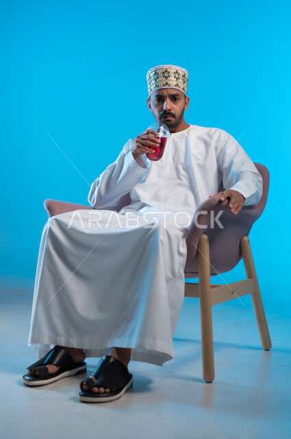 مفهوم عدم الرضا والقبول، النظر الى الكاميرا بإيماءات الغضب و الاستياء, محاولة التخفيف من حرارة الصيف بالمشروب المنعش، بورتريه لرجل عربي خليجي عماني مبتسم يرتدي اللكمة و الدشداشة يجلس على كرسي مريح بقوم بشرب العصير الطبيعي البارد, خلفية زرقاء