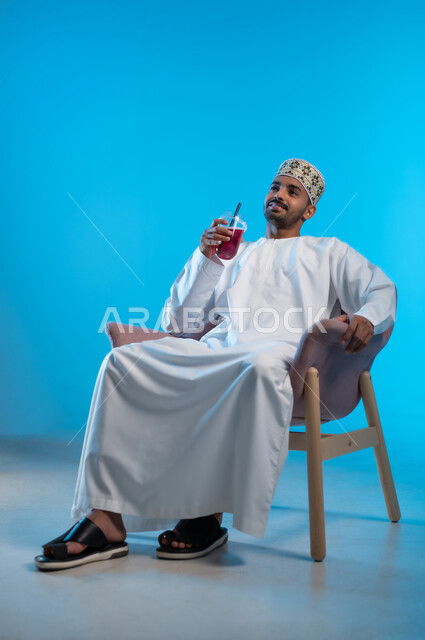 محاولة التخفيف من حرارة الصيف بالمشروب المنعش، الاستمتاع بالمشروبات المفيدة اللذيذة، بورتريه لرجل عربي خليجي عماني مبتسم يرتدي اللكمة و الدشداشة يجلس على كرسي مريح ويستمتع بشرب العصير الطبيعي البارد بإيماءات السرور والسعادة, خلفية زرقاء